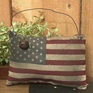 May include: A rectangular, patriotic-themed pillow with a distressed American flag design. The pillow features red and white stripes and a blue field with white stars. A small, brown bell hangs from a wire attached to the pillow.