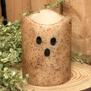 May include: A tan, cylindrical candle with a ghost face design. The candle has three black dots for eyes and mouth. The candle is speckled with brown spots and sits on a wooden slice with greenery.