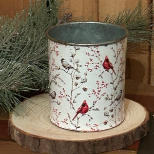 May include: A cylindrical metal container with a white background, adorned with illustrations of red cardinals and small birds perched on branches with red berries. The container sits on a round wooden slice.