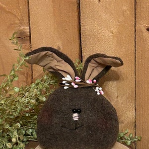 May include: A brown felt bunny with a pink and white beaded flower crown. The bunny is sitting on a wooden slice.