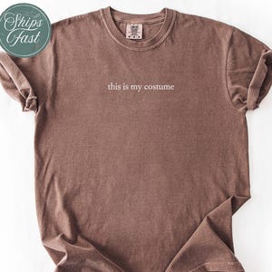 May include: Espresso-colored t-shirt with the text "this is my costume" in a simple, white font. The shirt has a vintage, washed look and rolled-up sleeves. A teal sticker with the words "Ships Fast" is in the upper left corner.