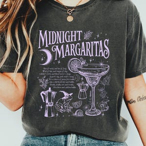 May include: Dark gray t-shirt with a purple graphic that reads "Midnight Margaritas." The design includes a cocktail glass, a crescent moon, and other illustrations. The shirt is worn with blue jeans and white sneakers.