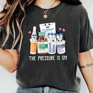 Puede incluir: Una camiseta gris oscuro con un gr&aacute;fico de suministros m&eacute;dicos de dibujos animados y el texto "THE PRESSURE IS ON".