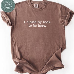 Puede incluir: Camiseta marrón con el texto "I closed my book to be here." La camiseta tiene cuello redondo y mangas cortas. Una pegatina verde y blanca "Ships Fast" está en la esquina superior izquierda.