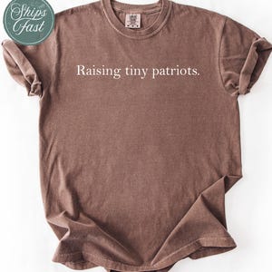 May include: A brown t-shirt with the text "Raising tiny patriots" in white. The shirt has a crew neck and short sleeves. The image also includes a green and white sticker that says "Ships fast".