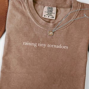 May include: A brown t-shirt with the text "raising tiny tornadoes" printed on the front. The shirt is folded and laid flat on a white surface.