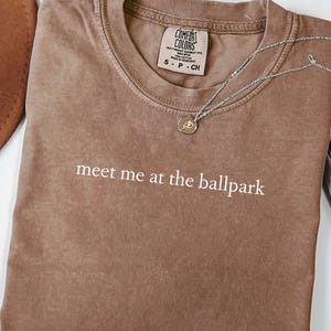 Puede incluir: Una camiseta marrón con el texto "meet me at the ballpark" en letras blancas. La camiseta está doblada y colocada sobre una superficie blanca. La camiseta es de estilo unisex.