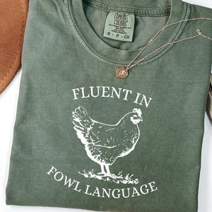 May include: A green t-shirt with the text "Fluent in Fowl Language" and a white silhouette of a chicken. The shirt is made of a comfortable, soft fabric.