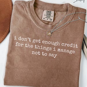 Puede incluir: Camiseta marr&oacute;n con la frase blanca "i don't get enough credit for the things i manage not to say". Un collar plateado con colgante descansa sobre la camiseta. La camiseta tiene una etiqueta Comfort Colors.