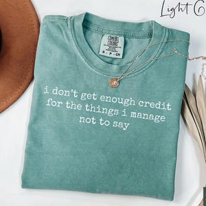 Puede incluir: Una camiseta gris claro con el texto "i don't get enough credit for the things i manage not to say" en blanco. Un collar dorado con un colgante est&aacute; drapeado sobre la camiseta. La camiseta est&aacute; doblada y se muestra con un sombrero y plantas secas decorativas.
