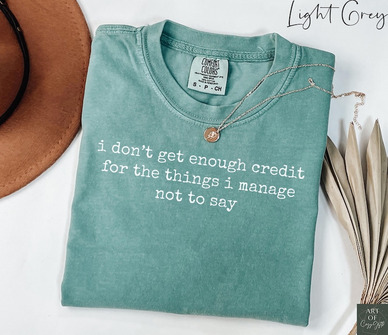 Puede incluir: Una camiseta gris claro con el texto "i don't get enough credit for the things i manage not to say" en blanco. Un collar dorado con un colgante est&aacute; drapeado sobre la camiseta. La camiseta est&aacute; doblada y se muestra con un sombrero y plantas secas decorativas.