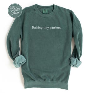 May include: A dark teal sweatshirt with the phrase "Raising tiny patriots" in white lettering. The long-sleeved crewneck shirt has rolled-up cuffs and a round "Ships Fast" sticker in the upper left corner. The garment is displayed on a white background.