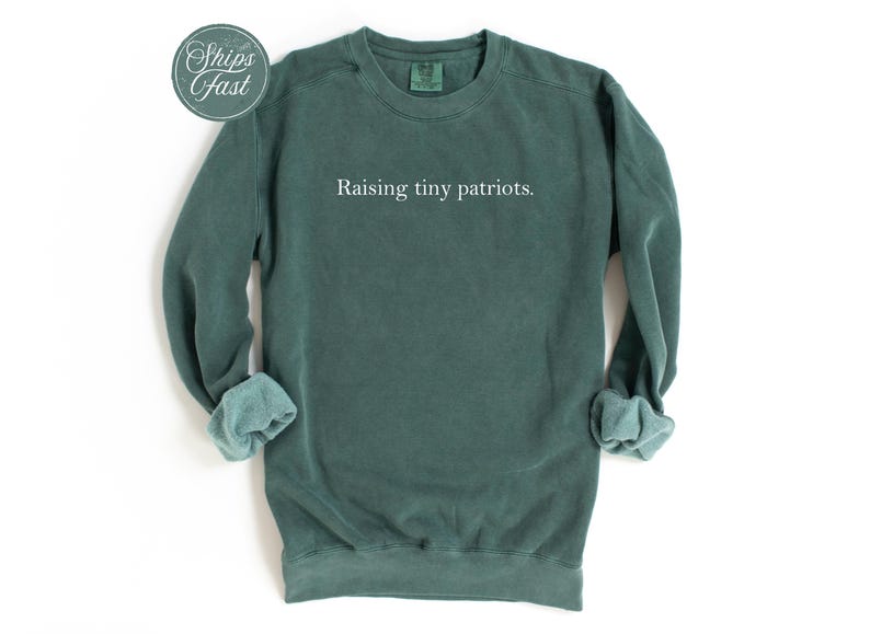 May include: A dark teal sweatshirt with the phrase "Raising tiny patriots" in white lettering. The long-sleeved crewneck shirt has rolled-up cuffs and a round "Ships Fast" sticker in the upper left corner. The garment is displayed on a white background.
