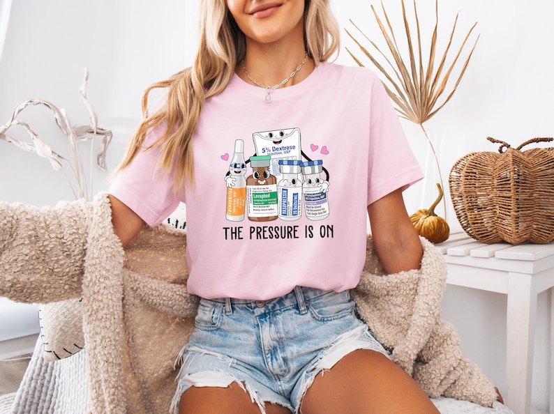 Puede incluir: Una camiseta rosa con un gr&aacute;fico de suministros m&eacute;dicos de dibujos animados y el texto "The Pressure Is On".