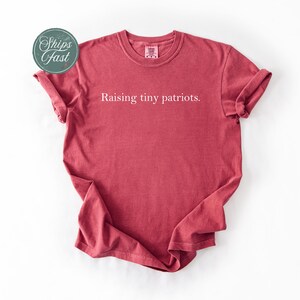 May include: A red t-shirt with the text "Raising tiny patriots." The shirt has rolled-up sleeves and a crew neck. A circular sticker with the words "Ships Fast" is in the upper left corner.