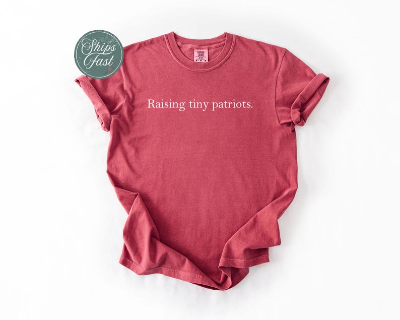 May include: A red t-shirt with the text "Raising tiny patriots." The shirt has rolled-up sleeves and a crew neck. A circular sticker with the words "Ships Fast" is in the upper left corner.