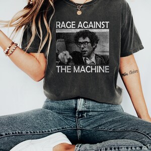 May include: A dark grey t-shirt with a black and white image of a man wearing glasses and a tie. The text "RAGE AGAINST THE MACHINE" is printed on the shirt.