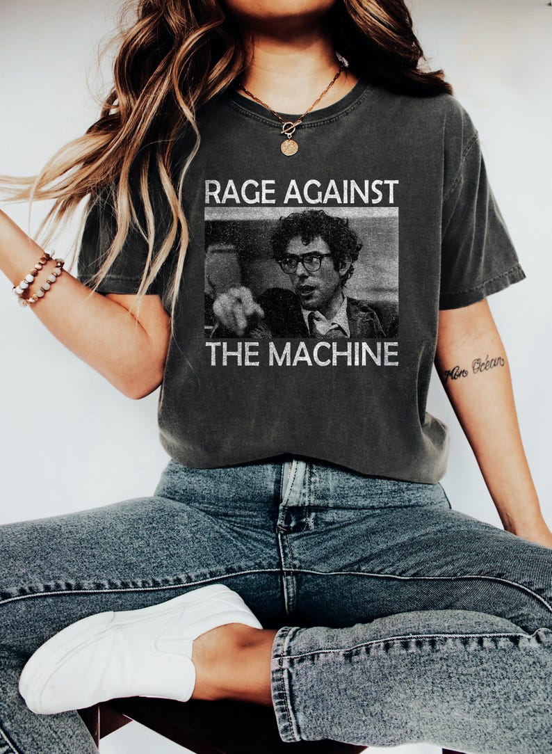 May include: A dark grey t-shirt with a black and white image of a man wearing glasses and a tie. The text "RAGE AGAINST THE MACHINE" is printed on the shirt.