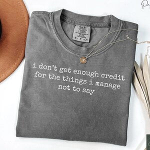 Puede incluir: Una camiseta gris Comfort Colors con el texto "i don't get enough credit for the things i manage not to say" en blanco. Un collar dorado con un colgante est&aacute; sobre la camiseta. Un sombrero marr&oacute;n est&aacute; en el fondo.