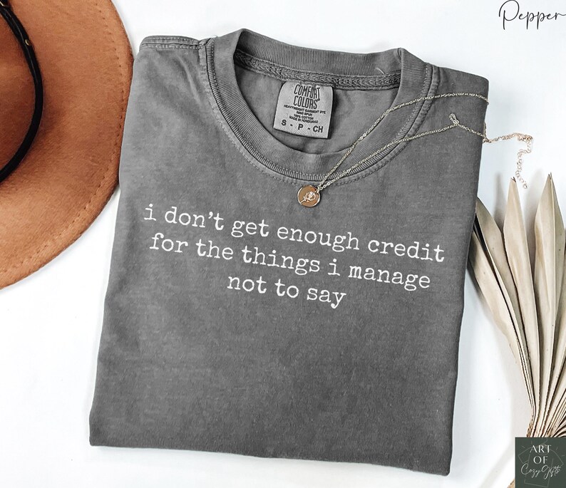 Puede incluir: Una camiseta gris Comfort Colors con el texto "i don't get enough credit for the things i manage not to say" en blanco. Un collar dorado con un colgante est&aacute; sobre la camiseta. Un sombrero marr&oacute;n est&aacute; en el fondo.