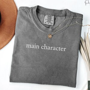 May include: A gray t-shirt with the text "main character" printed in white on the front. The shirt is made of a soft, comfortable material and has a relaxed fit.