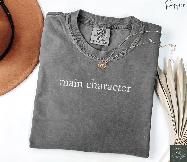 May include: A gray t-shirt with the text "main character" printed in white on the front. The shirt is made of a soft, comfortable material and has a relaxed fit.