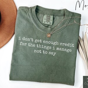 Puede incluir: Una camiseta verde musgo con el texto "i don't get enough credit for the things i manage not to say" en blanco. Un collar dorado con un colgante descansa sobre la camiseta.