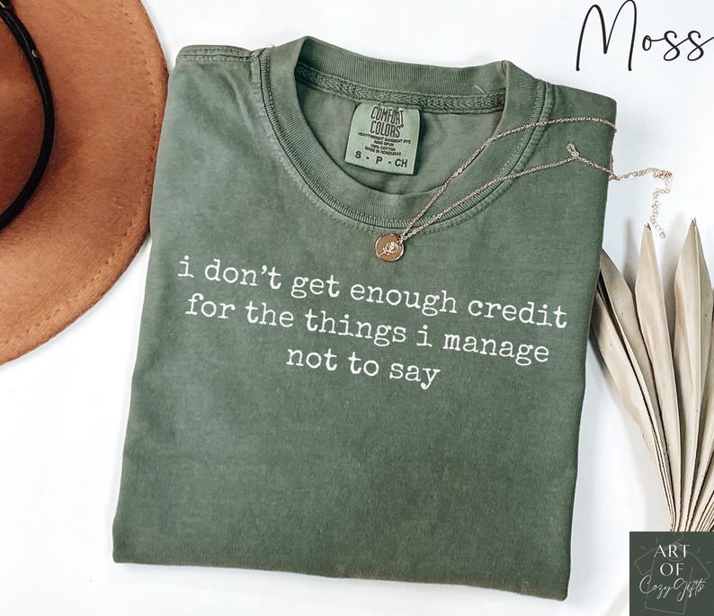 Puede incluir: Una camiseta verde musgo con el texto "i don't get enough credit for the things i manage not to say" en blanco. Un collar dorado con un colgante descansa sobre la camiseta.