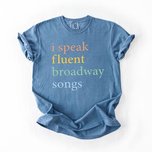 May include: A blue t-shirt with the text "i speak fluent broadway songs" printed on the front in rainbow colors.