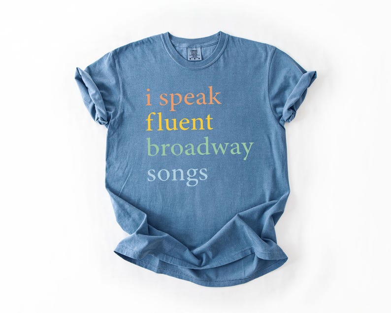 May include: A blue t-shirt with the text "i speak fluent broadway songs" printed on the front in rainbow colors.