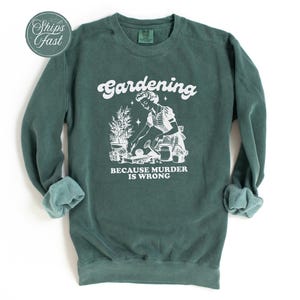 Peut inclure: Sweat-shirt vert fonc&eacute; avec texte et graphiques blancs. Le texte dit "Gardening" au-dessus d'une illustration d'une personne jardinant, avec les mots "BECAUSE MURDER IS WRONG" en dessous. Les manches sont retrouss&eacute;es.