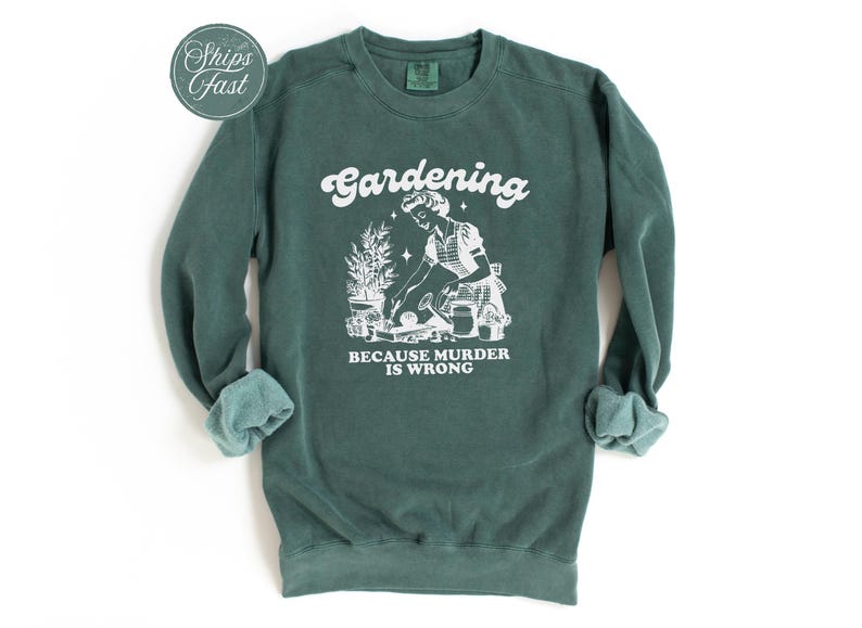 Peut inclure: Sweat-shirt vert fonc&eacute; avec texte et graphiques blancs. Le texte dit "Gardening" au-dessus d'une illustration d'une personne jardinant, avec les mots "BECAUSE MURDER IS WRONG" en dessous. Les manches sont retrouss&eacute;es.