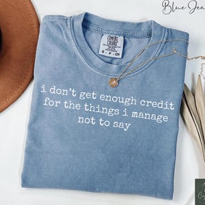 Puede incluir: Una camiseta azul Comfort Colors con el texto blanco "i don't get enough credit for the things i manage not to say". Un collar dorado con un colgante descansa sobre la camiseta.