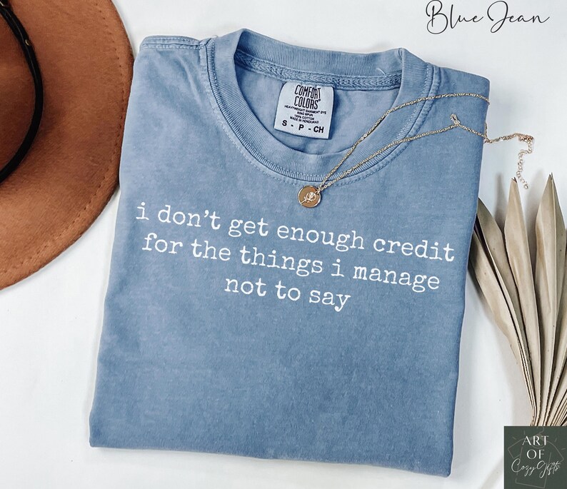 Puede incluir: Una camiseta azul Comfort Colors con el texto blanco "i don't get enough credit for the things i manage not to say". Un collar dorado con un colgante descansa sobre la camiseta.