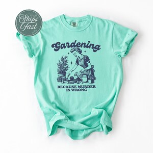 Peut inclure: T-shirt vert clair avec le mot "Gardening" au-dessus d'une illustration vintage d'une femme jardinant. Le texte "BECAUSE MURDER IS WRONG" est en dessous. Le t-shirt a des manches retrouss&eacute;es.