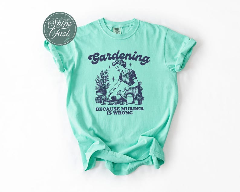 Peut inclure: T-shirt vert clair avec le mot "Gardening" au-dessus d'une illustration vintage d'une femme jardinant. Le texte "BECAUSE MURDER IS WRONG" est en dessous. Le t-shirt a des manches retrouss&eacute;es.