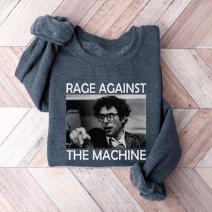 May include: A dark gray sweatshirt with the text "Rage Against The Machine" printed in white on the front. The text is centered on the sweatshirt and is surrounded by a black rectangle. A black and white image of a man with glasses is inside the rectangle.