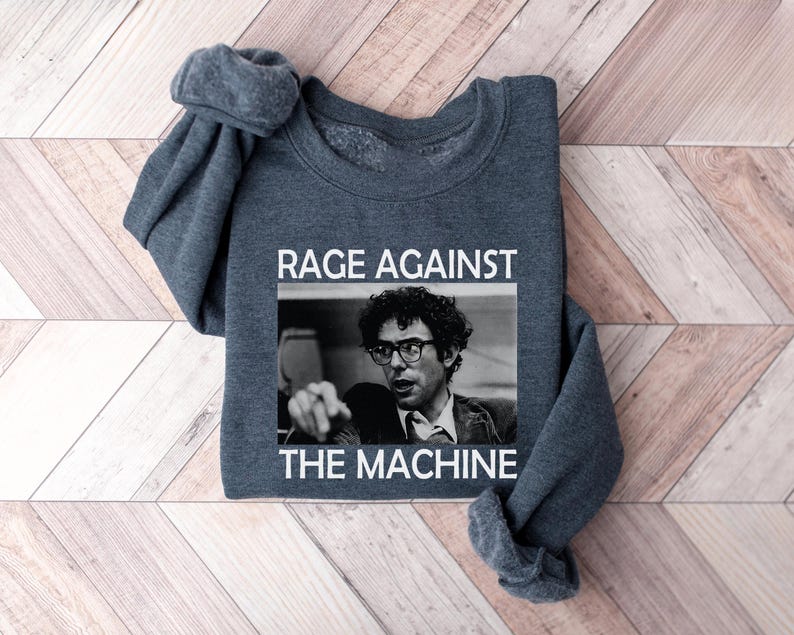 May include: A dark gray sweatshirt with the text "Rage Against The Machine" printed in white on the front. The text is centered on the sweatshirt and is surrounded by a black rectangle. A black and white image of a man with glasses is inside the rectangle.