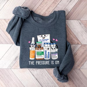 Puede incluir: Una sudadera gris oscuro con un gr&aacute;fico de suministros m&eacute;dicos, que incluye una bolsa de dextrosa, un vial de levotiroxina y dos viales de soluci&oacute;n salina. El gr&aacute;fico est&aacute; rodeado de corazones y el texto "The Pressure Is On".