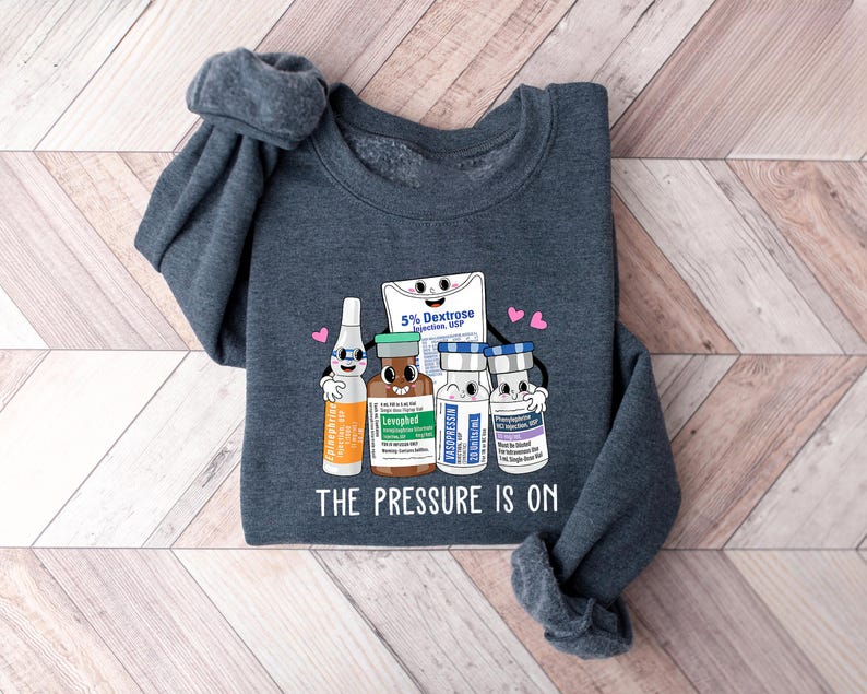 Puede incluir: Una sudadera gris oscuro con un gr&aacute;fico de suministros m&eacute;dicos, que incluye una bolsa de dextrosa, un vial de levotiroxina y dos viales de soluci&oacute;n salina. El gr&aacute;fico est&aacute; rodeado de corazones y el texto "The Pressure Is On".