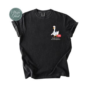 May include: Black t-shirt featuring a white cartoon goose wearing a stethoscope and carrying a red first aid kit. The text "honk if it's an emergency" is printed below the goose.