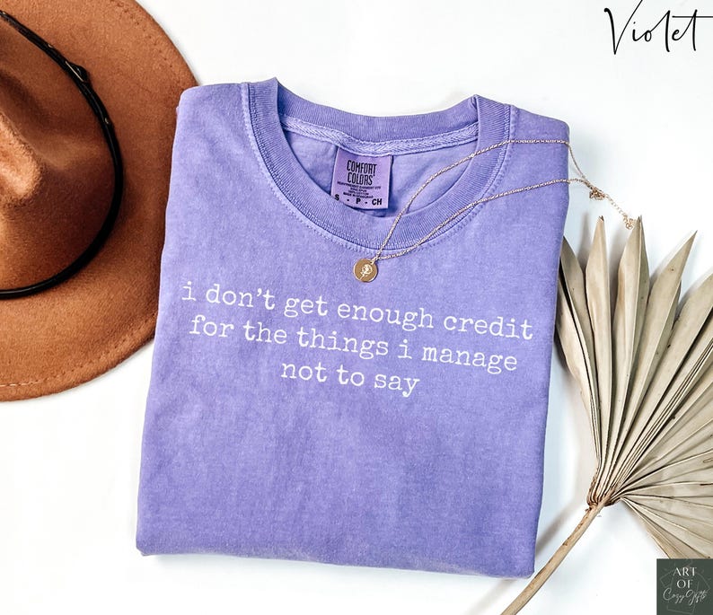 Puede incluir: Una camiseta de color violeta con el texto "i don't get enough credit for the things i manage not to say" en blanco. Un collar dorado con un colgante est&aacute; sobre la camiseta. Un sombrero marr&oacute;n y hojas de palma secas est&aacute;n en el fondo.