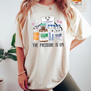 Puede incluir: Una camiseta blanca con un gr&aacute;fico de viales m&eacute;dicos de dibujos animados y una bolsa de soluci&oacute;n de dextrosa. El texto "THE PRESSURE IS ON" est&aacute; impreso debajo del gr&aacute;fico.
