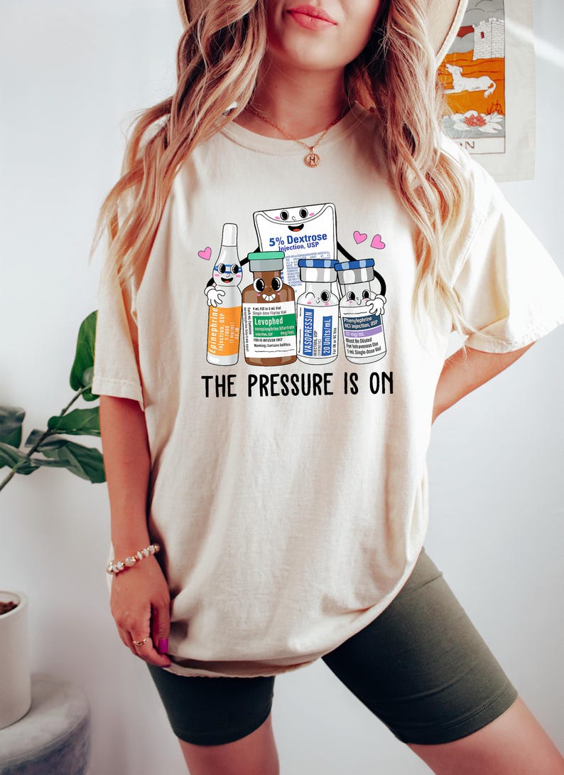 Puede incluir: Una camiseta blanca con un gr&aacute;fico de viales m&eacute;dicos de dibujos animados y una bolsa de soluci&oacute;n de dextrosa. El texto "THE PRESSURE IS ON" est&aacute; impreso debajo del gr&aacute;fico.
