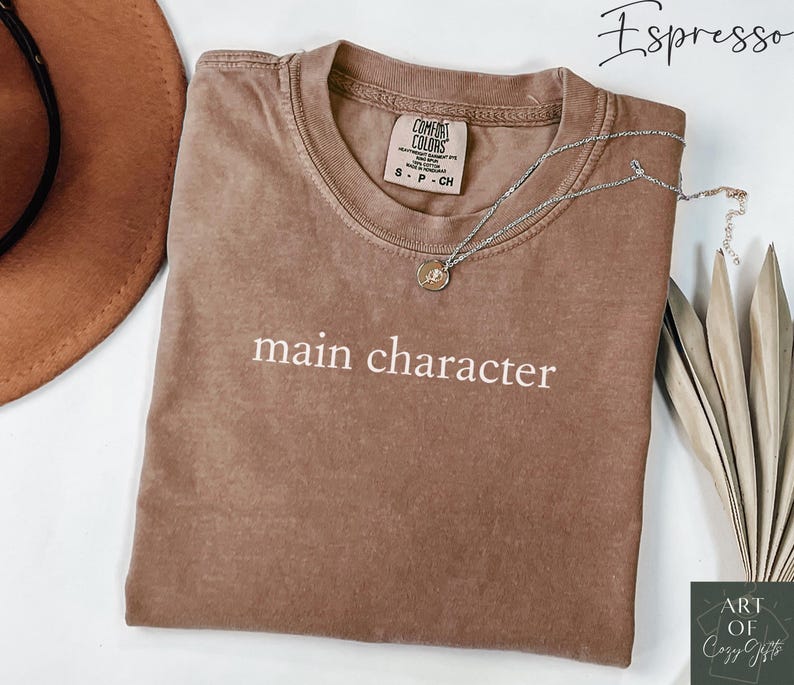 May include: A brown t-shirt with the text 'main character' in white. The shirt is folded and displayed on a white surface. The shirt is a comfort colors brand and has a tag that says 'S - P - CH'. There is a silver necklace with a coin pendant on the shirt. The background of the image is a white surface with a brown hat and a green plant.