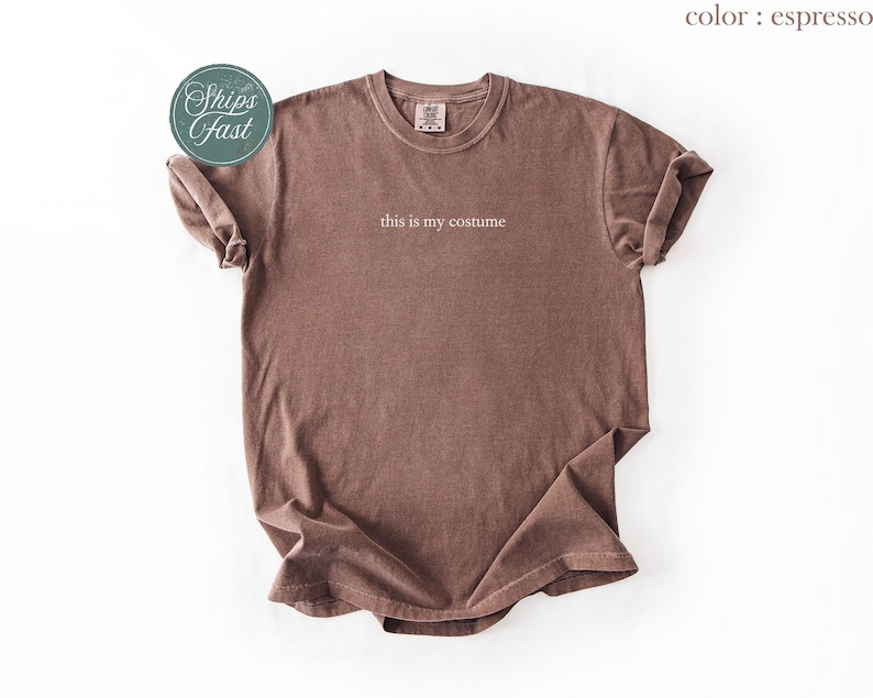 May include: Espresso-colored t-shirt with the text "this is my costume" in a simple, white font. The shirt has a vintage, washed look and rolled-up sleeves. A teal sticker with the words "Ships Fast" is in the upper left corner.