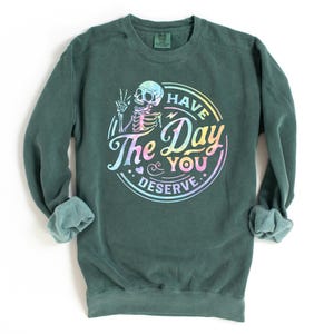 May include: A dark green sweatshirt with a rainbow-colored graphic of a skeleton holding a peace sign and the text "Have the Day You Deserve."