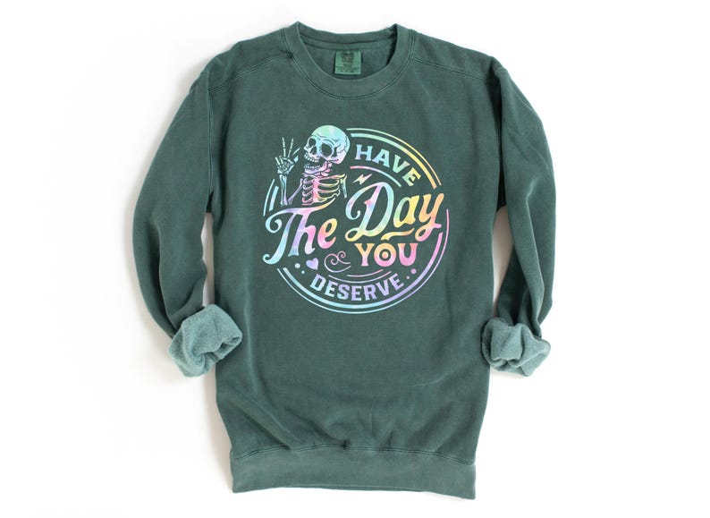 May include: A dark green sweatshirt with a rainbow-colored graphic of a skeleton holding a peace sign and the text "Have the Day You Deserve."