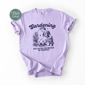 Peut inclure: Un t-shirt lilas clair avec le mot "Gardening" dans une police r&eacute;tro au-dessus d'une illustration d'une femme jardinant. Le texte "BECAUSE MURDER IS WRONG" est en dessous. Le t-shirt a des manches retrouss&eacute;es.