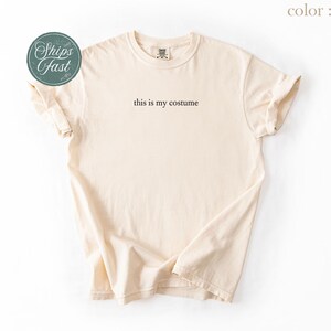 May include: Ivory-colored t-shirt with the phrase "this is my costume" printed in black. The shirt is folded, showing the sleeves and neckline. A round green sticker with the words "Ships Fast" is in the upper left corner.